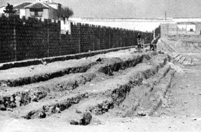 Fotografía en blanco y negro de las gradas de tierra en construcción junto al muro perimetral.