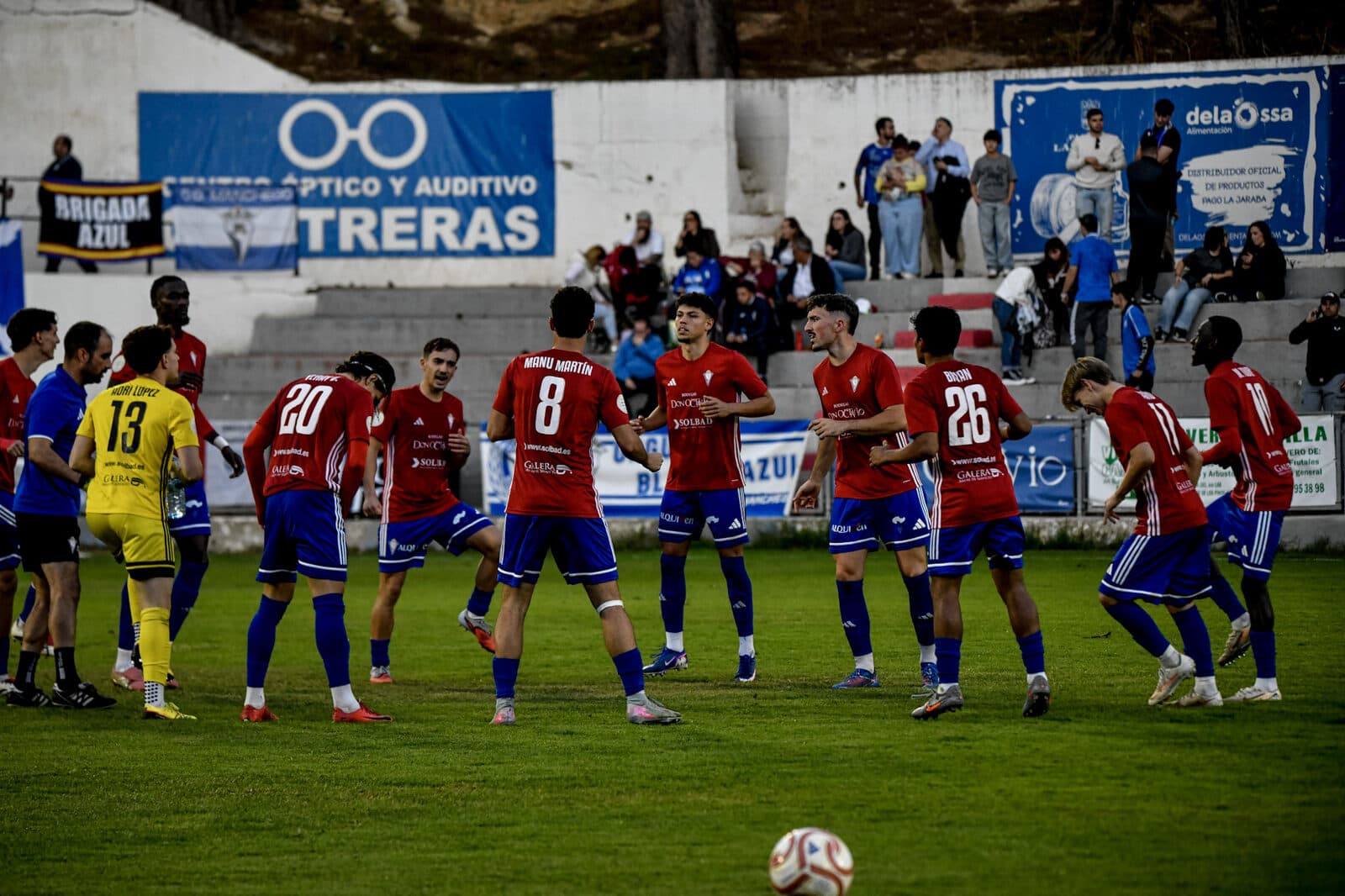 Jugadores del CP Villarrobledo, con camiseta roja y pantalón azul, sobre el césped del Municipal ante la grada.